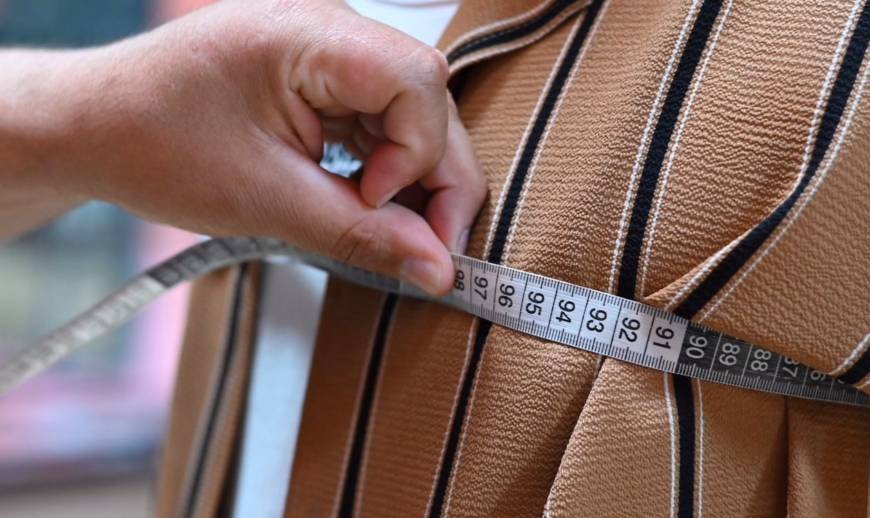 Close-up of a hand holding a soft measuring tape against a brown striped fabric sample, checking the dimensions of a product sample before placing a first bulk order.