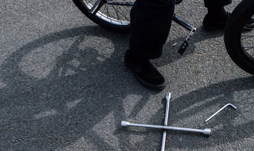A cyclist crouched beside a bike on asphalt with hex keys and socket tools laid out on the ground, showing the tools and hands-on setup used for a home disc brake adjustment.
