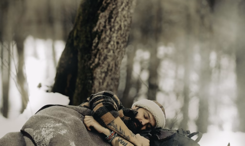 Camper resting under a thick insulating blanket beside a sleeping bag and pack in a snow-covered winter forest, illustrating how sleeping bag temperature ratings relate to overnight warmth in cold conditions