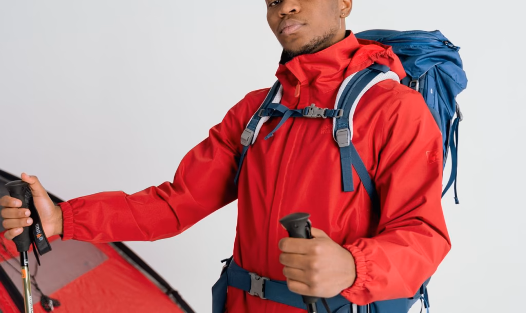 Hiker in a red windbreaker holding a pair of trekking poles at his sides, standing next to a pitched tent, illustrating correct trekking pole fit and length for hiking
